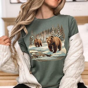 May include: A green t-shirt with a graphic print of two brown bears walking through a snowy forest scene. The bears are walking on a path with a small stream running through it. The forest is covered in snow and there are trees on both sides of the path.