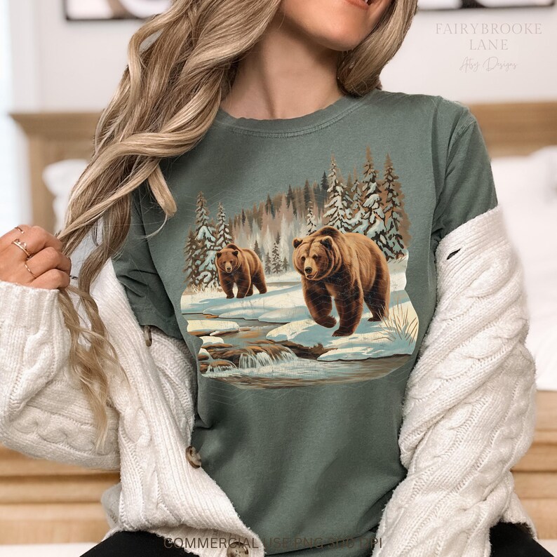 May include: A green t-shirt with a graphic print of two brown bears walking through a snowy forest scene. The bears are walking on a path with a small stream running through it. The forest is covered in snow and there are trees on both sides of the path.