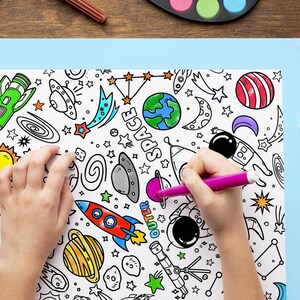 May include: A child's hands coloring a space-themed coloring page with a pink marker. The page features planets, rockets, stars, and the word "SPACE". Watercolor paints and markers are on the table.
