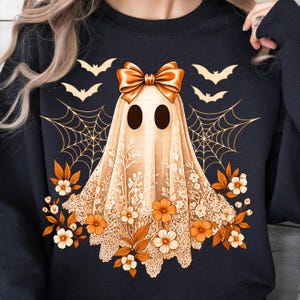 May include: A navy blue sweatshirt featuring a ghost design with a bow, lace veil, and floral accents. The ghost has two black eyes and is surrounded by bats and spiderwebs in a Halloween theme.
