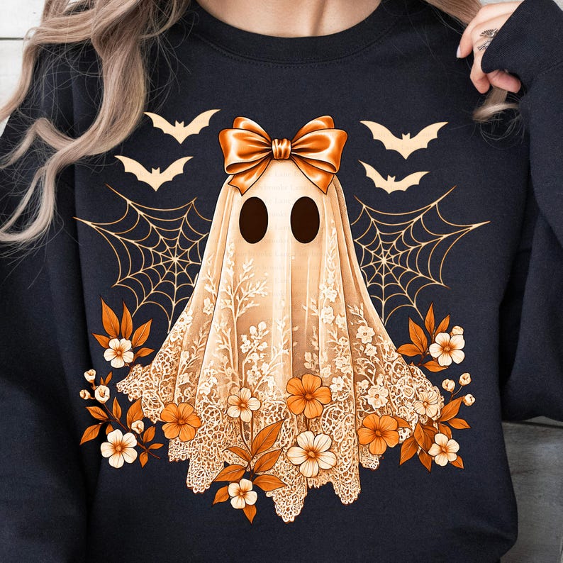 May include: A navy blue sweatshirt featuring a ghost design with a bow, lace veil, and floral accents. The ghost has two black eyes and is surrounded by bats and spiderwebs in a Halloween theme.