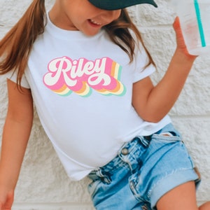 May include: A white t-shirt with a retro-style rainbow design featuring the name "Riley" in pink.