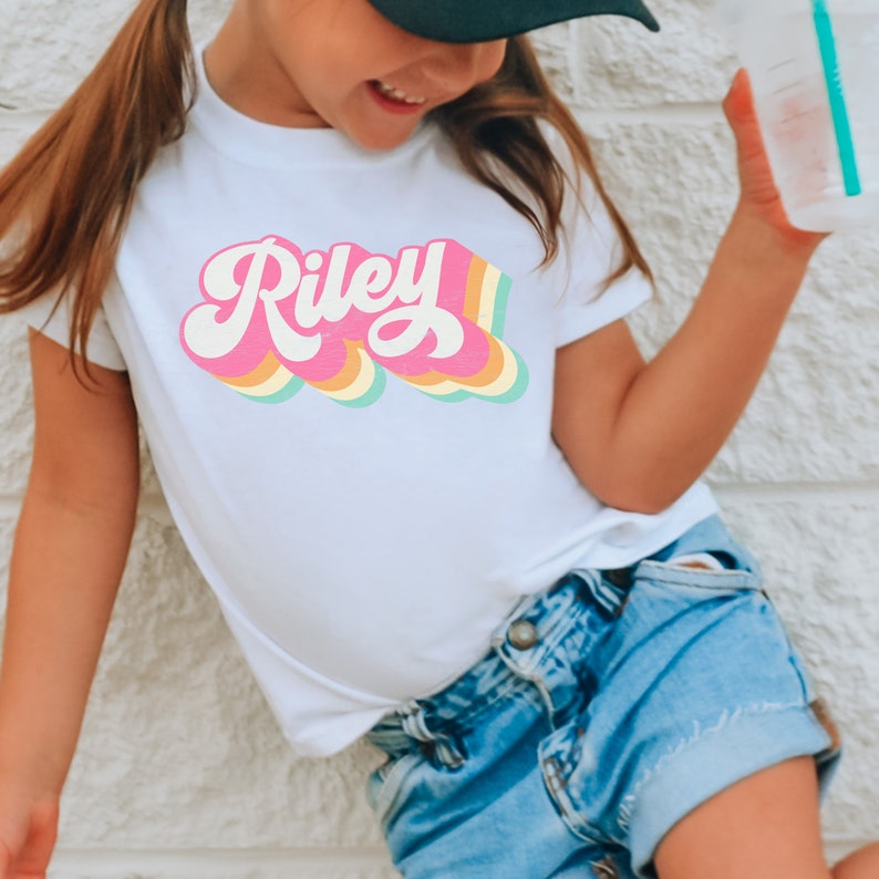 May include: A white t-shirt with a retro-style rainbow design featuring the name "Riley" in pink.