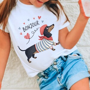 May include: White t-shirt featuring a cartoon dachshund wearing a red beret, scarf, and striped shirt. The word "BONJOUR" is above the dog, with heart and swirl graphics. The shirt is paired with blue denim shorts.