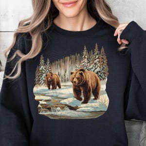 May include: A dark blue sweatshirt with a graphic print of two brown bears walking through a snowy forest scene. The bears are walking along a stream with a waterfall in the background.