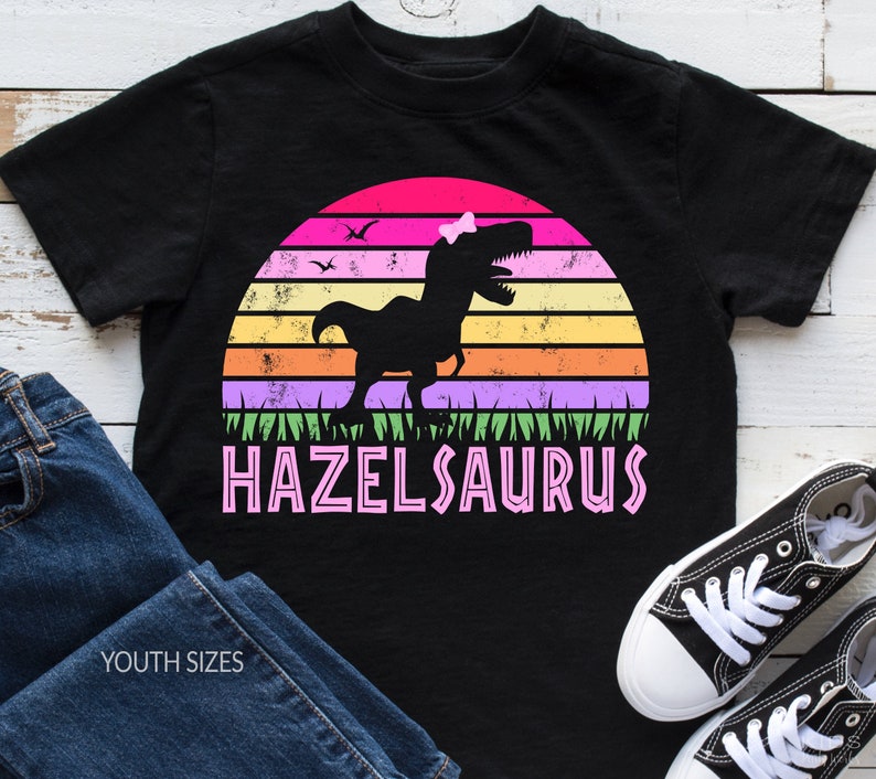 May include: Black youth t-shirt with a retro sunset graphic featuring a pink dinosaur with a bow. The text "HAZELSAURUS" is printed in pink below the dinosaur.