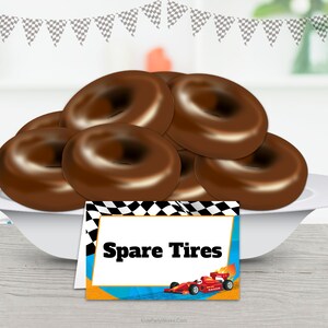 Puede incluir: Un plato blanco contiene varias rosquillas cubiertas de chocolate, que parecen neum&aacute;ticos de repuesto. Una tarjeta decorativa al frente dice "Spare Tires" con una ilustraci&oacute;n de un coche de carreras, ideal para una fiesta tem&aacute;tica de carreras. Guirnalda de banderas a cuadros en el fondo.