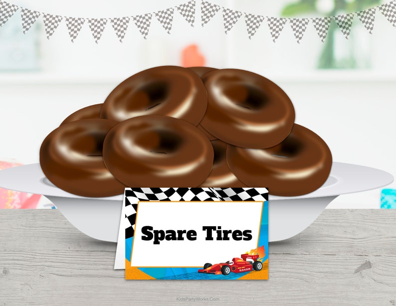 Puede incluir: Un plato blanco contiene varias rosquillas cubiertas de chocolate, que parecen neum&aacute;ticos de repuesto. Una tarjeta decorativa al frente dice "Spare Tires" con una ilustraci&oacute;n de un coche de carreras, ideal para una fiesta tem&aacute;tica de carreras. Guirnalda de banderas a cuadros en el fondo.