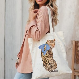 Puede incluir: Una bolsa de lona blanca con un dise&ntilde;o de estampado de leopardo y un lazo azul. La bolsa tiene el texto "Kids Party Works Design Studio" impreso en ella.