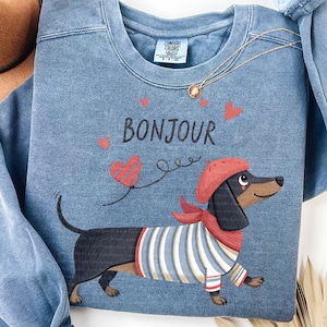 May include: Blue sweatshirt with a graphic of a dachshund wearing a red beret and scarf, and a striped shirt. The word "Bonjour" is printed above the dog, with heart accents.