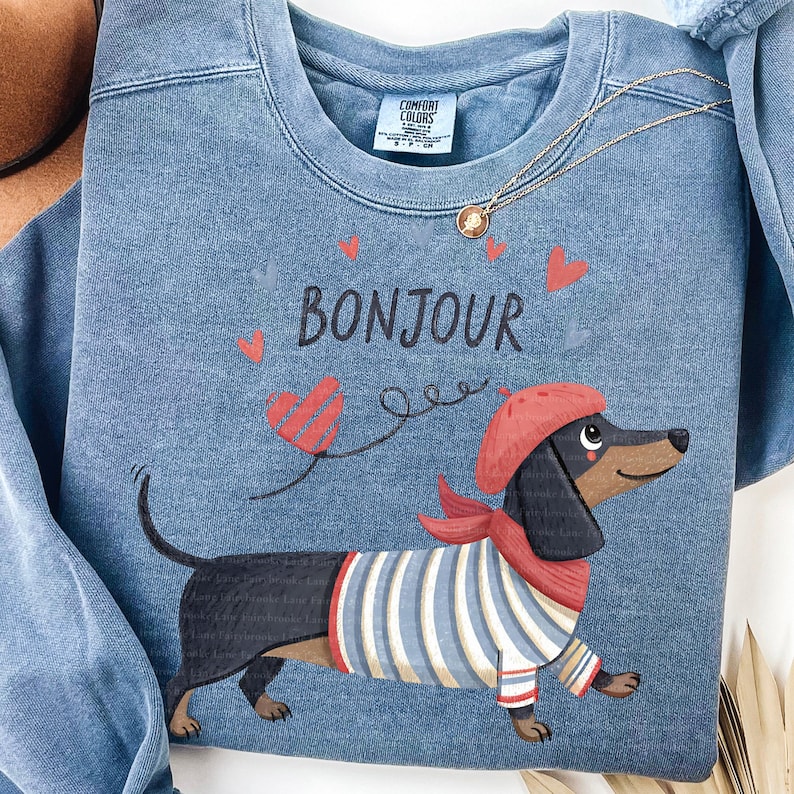 May include: Blue sweatshirt with a graphic of a dachshund wearing a red beret and scarf, and a striped shirt. The word "Bonjour" is printed above the dog, with heart accents.