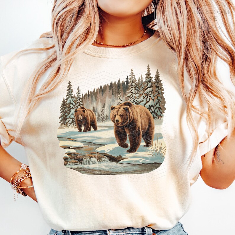 May include: A white t-shirt with a graphic of two brown bears walking through a snowy forest. The bears are walking on a path with a stream running through it. The forest is covered in snow and there are trees on both sides of the path.