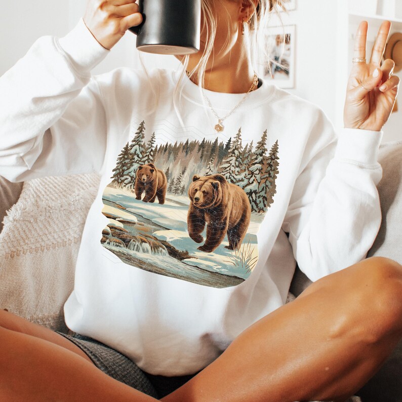 May include: White sweatshirt with a graphic print of two brown bears walking through a snowy forest. The bears are walking on a path with a stream running through it.