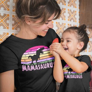 May include: A mother and daughter wearing matching black t-shirts with a pink dinosaur graphic and the text "Mamasaurus" and "Misaurus".