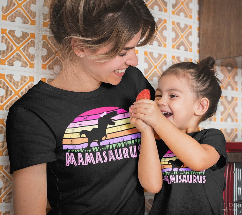 May include: A mother and daughter wearing matching black t-shirts with a pink dinosaur graphic and the text "Mamasaurus" and "Misaurus".