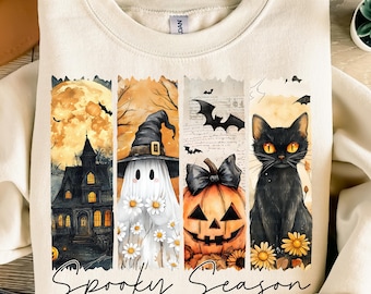 May include: Cream-colored sweatshirt with a Halloween-themed graphic. The design features a haunted house, a ghost wearing a witch hat, a jack-o'-lantern, and a black cat. The words "Spooky Season" are written in a script font.