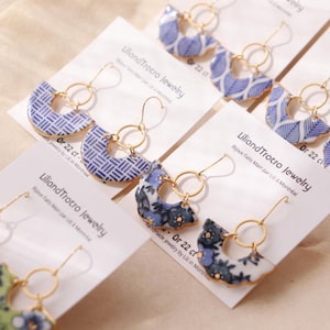 May include: Several pairs of dangle earrings featuring semi-circular patterned charms in blue, white, and green floral or geometric designs. Each charm is attached to a gold-toned circular hoop and hook, displayed on white cards labelled LiliandTrotro Jewelry.
