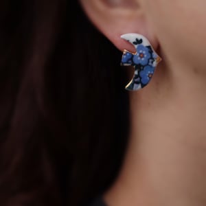 Peut inclure: Une boucle d'oreille en céramique en forme d'oiseau en plein vol, présentant une base blanche avec un motif floral bleu, des accents bleu foncé et un fin liseré métallique doré sur les bords, fixée au lobe par une tige.