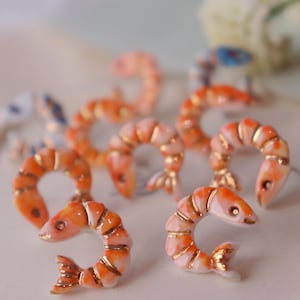 Op de afbeelding: Close-up van verschillende oranje en witte garnaalvormige decoratieve objecten met gouden accenten. De garnalen zijn gerangschikt op een wit oppervlak. De objecten hebben een glanzende afwerking en zijn gedetailleerd met gouden randen.