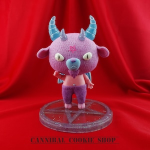 Puede incluir: Una figurita de diablo rosa y azul con cuernos y un pentagrama en la frente. La figurita está de pie sobre una base con forma de pentagrama con purpurina.