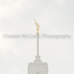 May include: A white spire with a golden statue of an angel holding a trumpet on top. The statue is against a white cloudy sky.