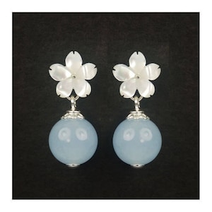 May include: A pair of earrings featuring white flower-shaped tops and light blue spherical drops. The earrings have silver-colored metal accents. The background is a solid black.