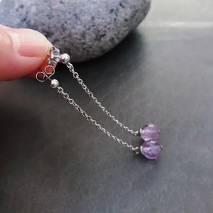 Könnte beinhalten: Silberne Kettenohrringe mit zwei kleinen, runden, lila Amethystperlen. Die Ohrringe haben ein Stecker- und Ketten-Design, wobei die Amethystperlen unten baumeln. Der Hintergrund ist grau.