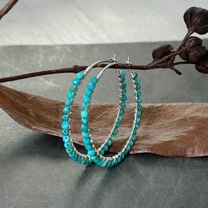 May include: Turquoise beaded hoop earrings. The earrings feature small, round turquoise beads strung on silver wire. The hoops are displayed on a brown leaf against a dark grey background.