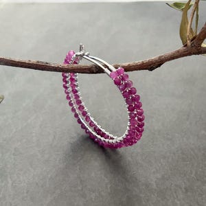 Puede incluir: Un pendiente de aro circular con un diseño de doble capa de pequeñas cuentas facetadas de color magenta, envueltas firmemente alrededor de una estructura de alambre de metal fino en tono plateado, creando una apariencia texturizada y vibrante.
