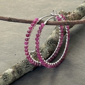 Op de afbeelding: Een paar oorringen met kleine, gefacetteerde, donkerroze edelsteenkralen gewikkeld rond een dunne, zilverkleurige metalen draadstructuur. De ronde oorbellen zijn geconstrueerd met een doorlopend draadontwerp dat de kralen op hun plek houdt.