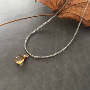 May include: A delicate necklace with a teardrop-shaped topaz pendant. The necklace is made of tiny, clear beads and is strung on a gold wire.