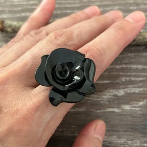Könnte beinhalten: Ein Ring mit einer großen, glänzend schwarzen Rose als Herzstück. Die Rose hat geschichtete, glatte Blütenblätter, die einen dreidimensionalen, skulpturalen Look erzeugen. Das schwarze Material hat eine reflektierende, polierte Oberfläche.