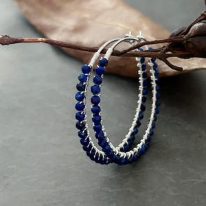 May include: A pair of hoop earrings with a silver wire design, embellished with small, round, deep blue beads. The earrings are set against a dark grey backdrop, with a brown twig and leaf.