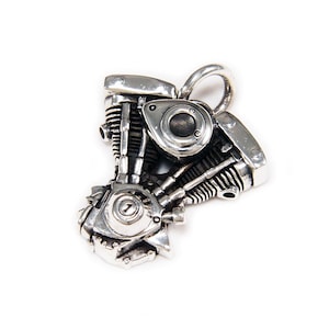 Può includere: Un ciondolo color argento a forma di motore di motocicletta. Il ciondolo presenta componenti meccanici dettagliati, tra cui cilindri, valvole e un elemento circolare centrale. Il design è intricato e realistico, con un anello per una catena.