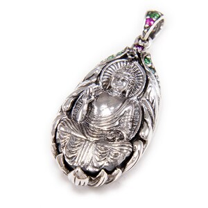 May include: Silver pendant featuring a detailed, ornate design of a seated Buddha with green and pink gemstones.