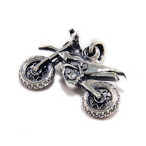 May include: A silver-toned metal pendant shaped like a dirt bike motorcycle. The charm features detailed wheels with spokes, a frame, handlebars, and a seat. A small circular jump ring is attached to the top for hanging on a chain.