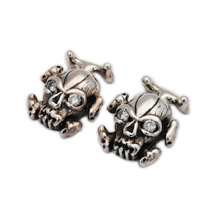 May include: A pair of silver-toned skull cufflinks. Each skull has a detailed design with visible teeth and sparkling crystal eyes. The cufflinks have a post-style backing for secure attachment to a shirt cuff.