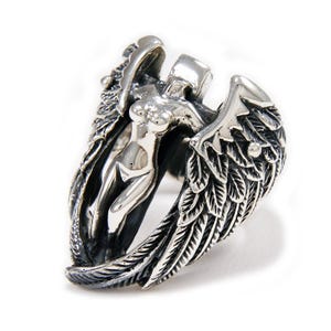 May include: A silver ring depicting a female form with wings. The ring has detailed feathering on the wings and a smooth, polished finish on the central figure. The ring is set against a white background.