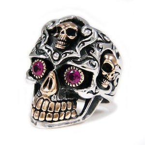 Puede incluir: Un anillo de calavera de plata con detalles intrincados, que presenta dos ojos rojo rubí y una calavera de oro en la parte superior. El anillo está adornado con volutas ornamentadas.