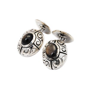 May include: A pair of silver cufflinks with a dark gray oval stone set in each. The cufflinks have an ornate design with swirls and scrolls.