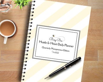 May include: A white and yellow striped daily planner with a black border and the text "Busy Bee Hustle & Heart Daily Planner Quarterly Mompreneur Edition Undated".