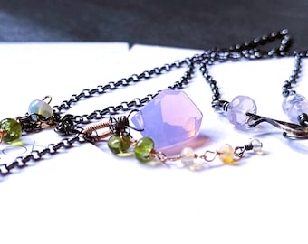 May include: Close-up of several necklaces with delicate chains and gemstone pendants. The necklaces feature a variety of stones, including opalescent, green, and lavender-colored gems. The chains are dark, and the overall aesthetic is elegant and handcrafted.