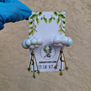 May include: A pair of iridescent white cloud earrings with gold star and crescent moon charms hanging from them. The earrings are on a white card with the text "GORGEPLUGS.COM" printed on it.