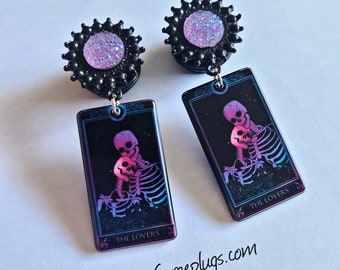 Puede incluir: Un par de dilatadores de oreja negros y morados con un diseño brillante rosa y blanco. Los dilatadores presentan un diseño de carta de tarot con el texto "The Lovers" y una pareja de esqueletos.