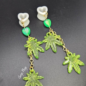 May include: A pair of white heart-shaped ear gauges with dangling green marijuana leaf charms and green heart beads. The charms are connected by a gold chain.