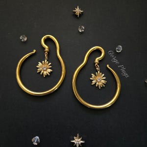 Può includere: Un paio di orecchini color oro dal design unico. Ogni orecchino presenta una forma curva e allungata con un pendente a margherita pendente. Il pendente a margherita ha una gemma centrale ed è circondato da delicati petali. Gli orecchini sono esposti su una superficie nera.