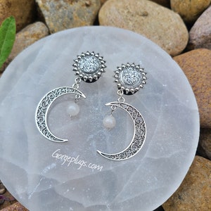 Könnte beinhalten: Ein Paar silberne Ohrdehner mit Mond- und Sonnenmotiv. Die Sonne ist mit glitzernden Strasssteinen verziert und der Mond hat einen baumelnden weißen Stein.