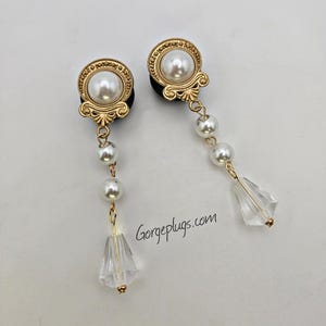 Puede incluir: Un par de dilatadores de oreja de tono dorado con un colgante de perla y cristal. Los dilatadores presentan un diseño decorativo con una perla en el centro. El colgante presenta una perla y una lágrima de cristal.