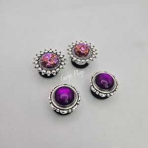 May include: Four decorative ear plugs with silver-colored, floral-shaped borders. Two plugs have a sparkling, glittery, pink and orange center. The other two plugs have a smooth, deep purple center. The text "Gorge Plugs" is visible.