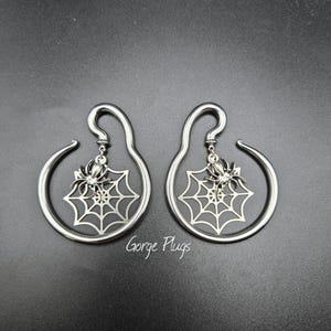 May include: Two silver-toned ear gauges with a spider and web design. Each gauge has a curved hook and a circular dangle. The spider is centered on a web. The text "Gorge Plugs" is at the bottom.